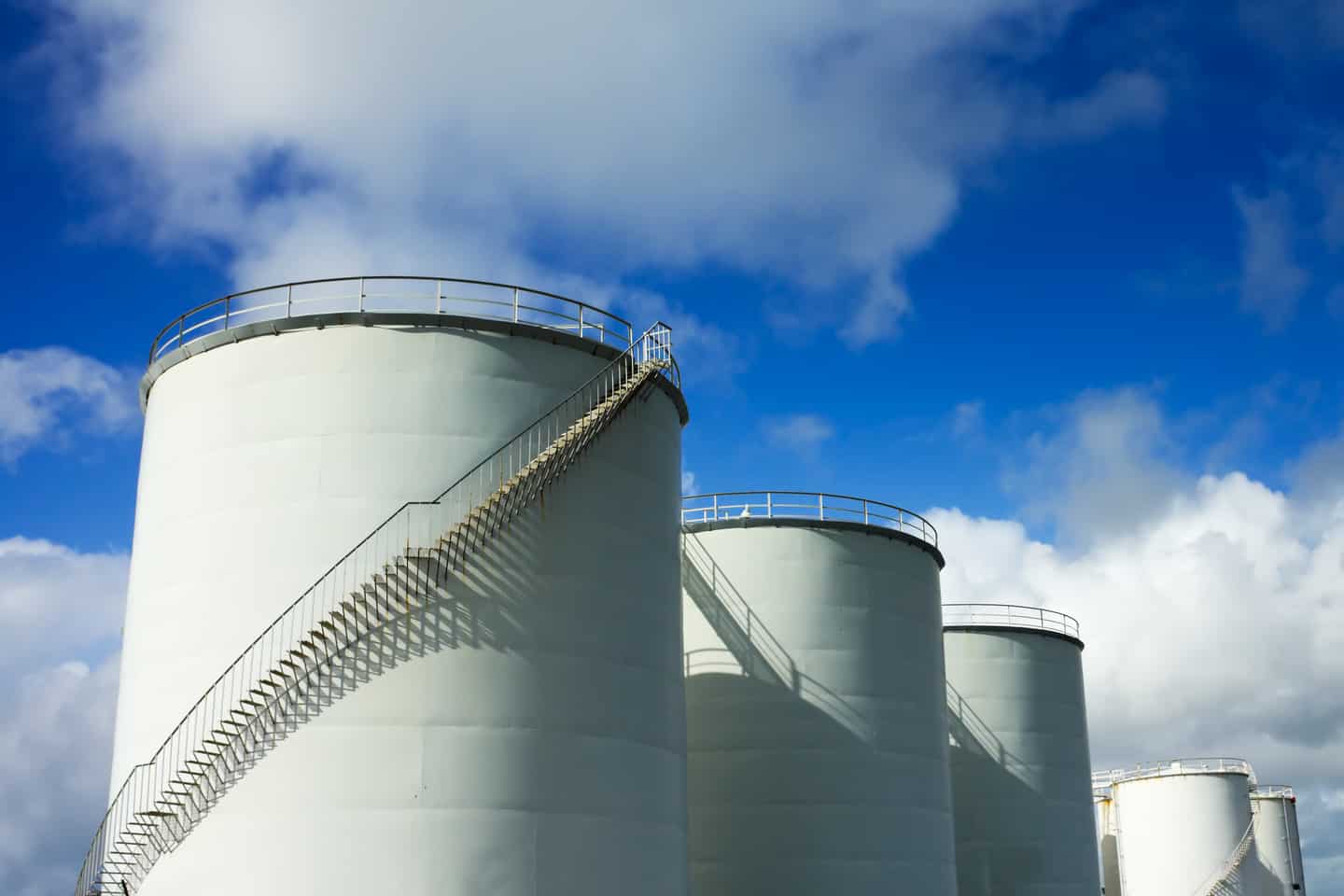 Multiple large white cylindrical storage tanks with spiral staircases under a bright blue sky with scattered clouds, likely used for storing liquids in an industrial facility.